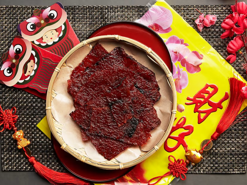 Chinese New Year bak kwa displayed on a bamboo plate with red tassels, lion envelopes, and festive ornaments.